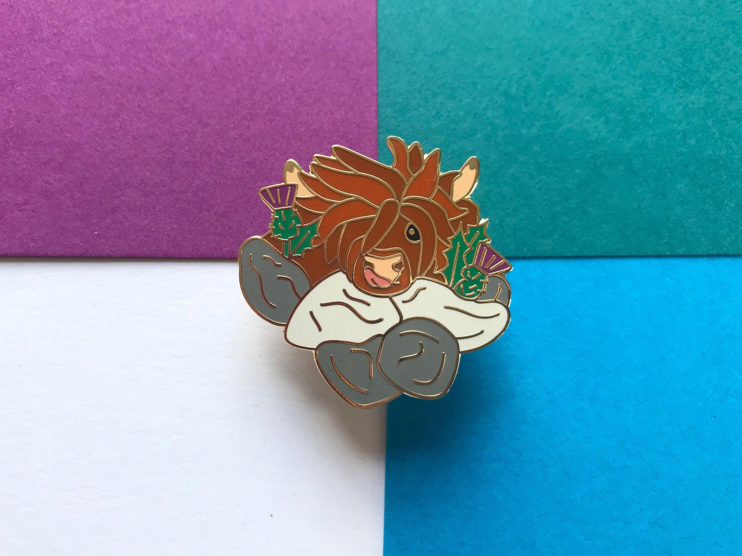 A photo of a funny highland cow enamel pin with thistles and a wall