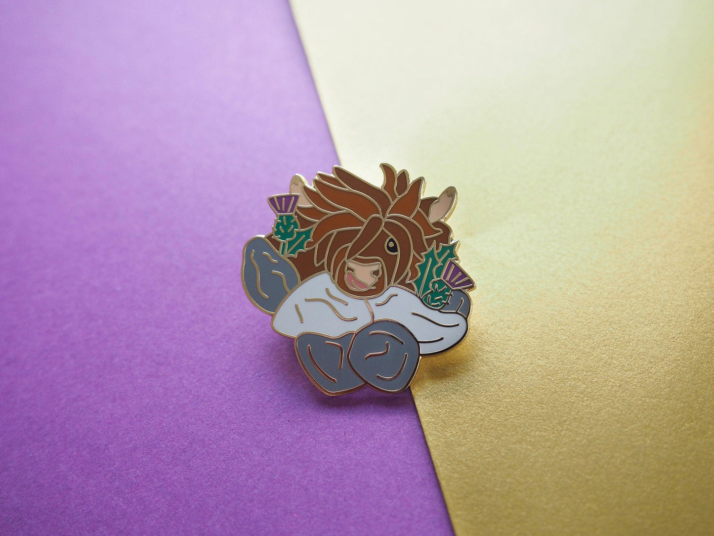 A photo of a funny highland cow enamel pin with thistles and a wall