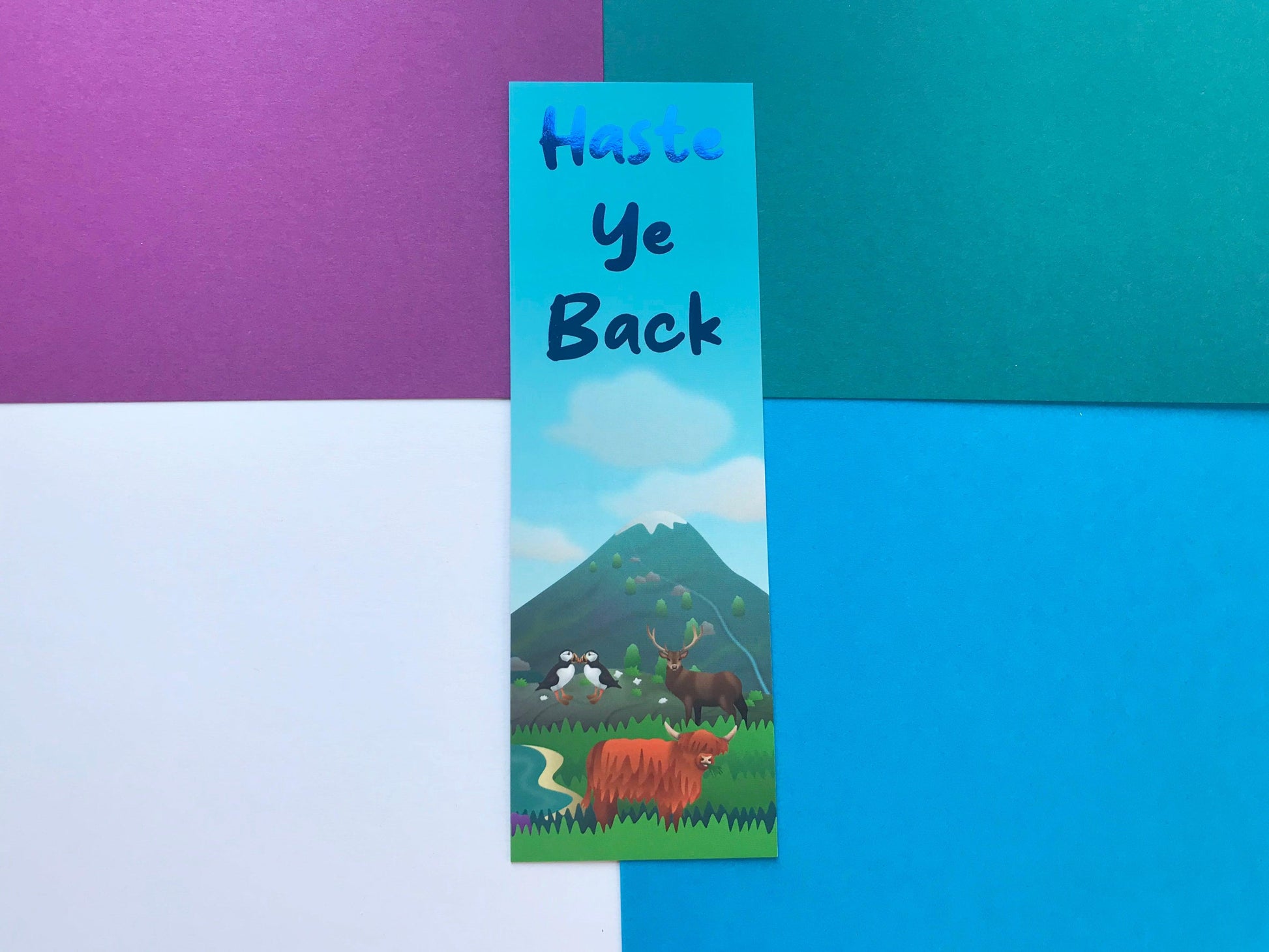 A photo of a bookmark that says "Haste ye Back" in blue foil letters, with a picture of a Scottish scene with animals