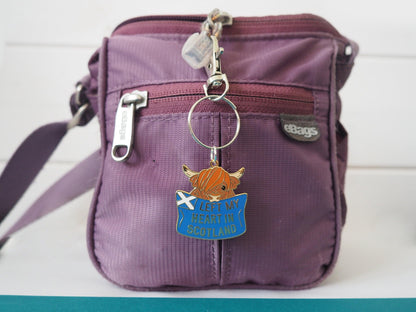 A photo of a purple bag with an enamel keychain that has a highland Cow holding a blue sign with the words "Left My Heart in Scotland" on it
