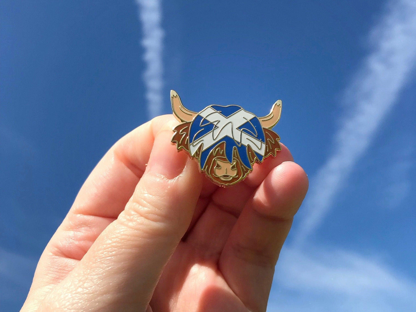 A photo of a blue sky with a Highland Cow Enamel Pin with a blue and white Saltire flag in its hair