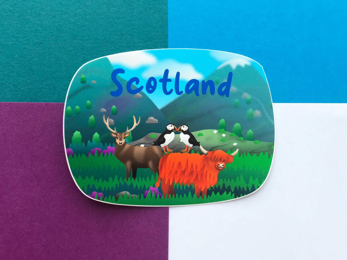 A photo of a vinyl Sticker with Scotland written on it in blue letters and a Scottish scene of highland cow, stag and 2 puffins