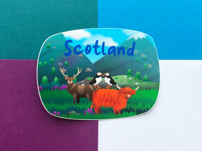 A photo of a vinyl Sticker with Scotland written on it in blue letters and a Scottish scene of highland cow, stag and 2 puffins