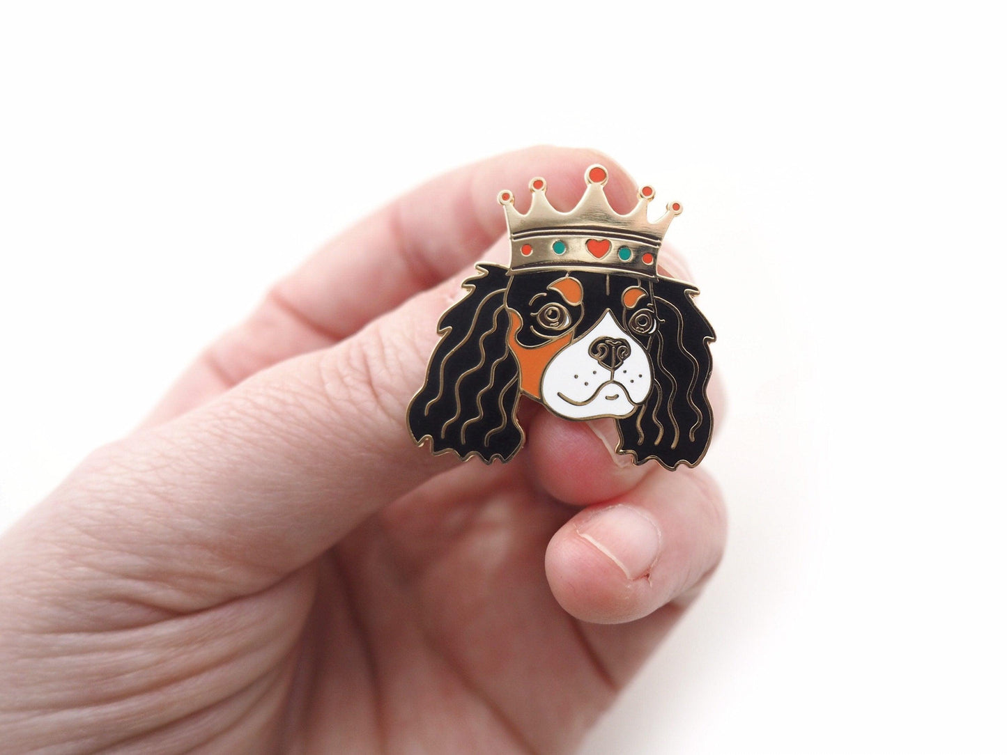 A photo of a hand holding a tri colour Cavaloer King Charles enamel Pin