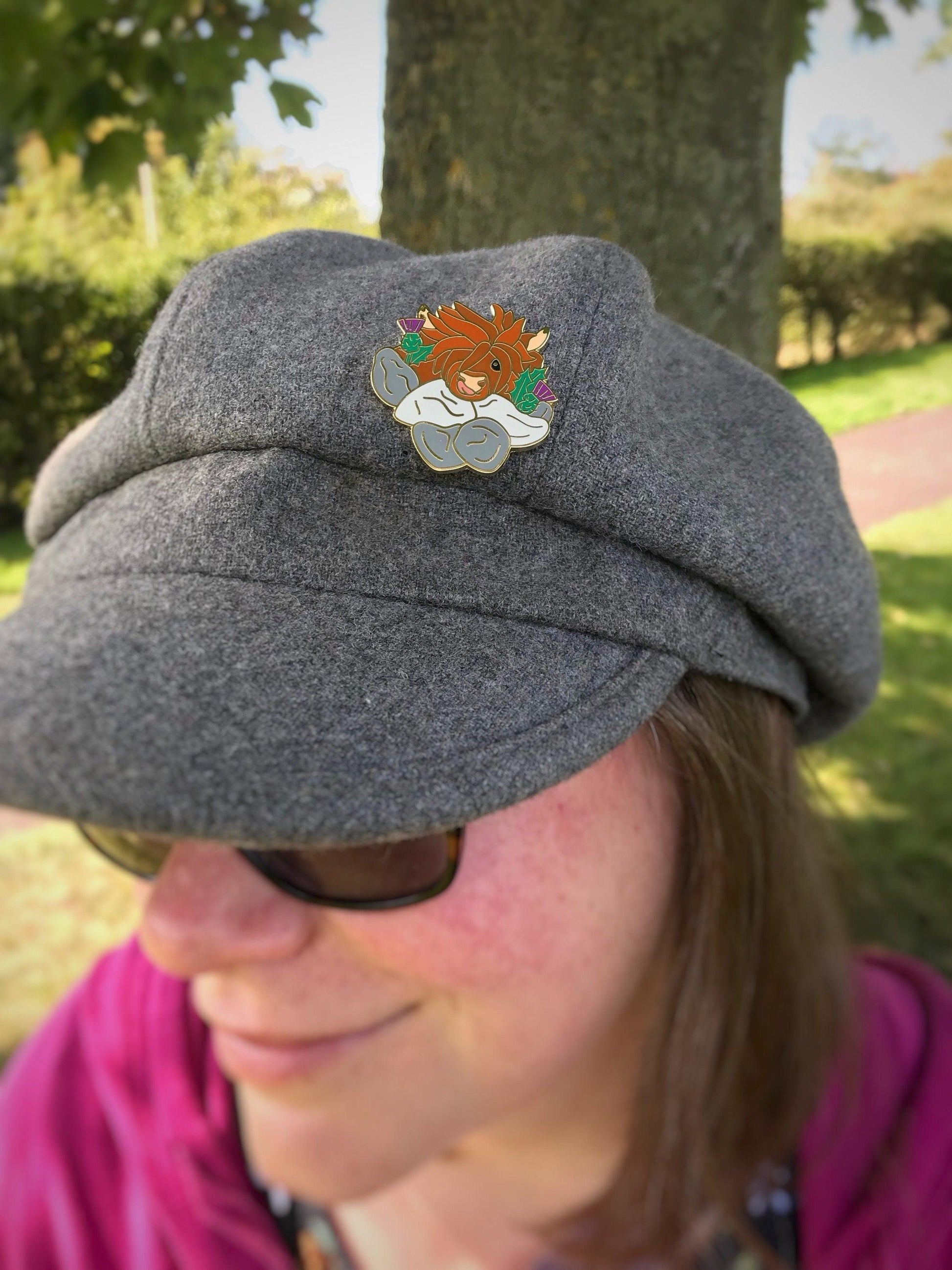 A photo of a funny highland cow enamel pin with thistles and a wall Pinned to a hat